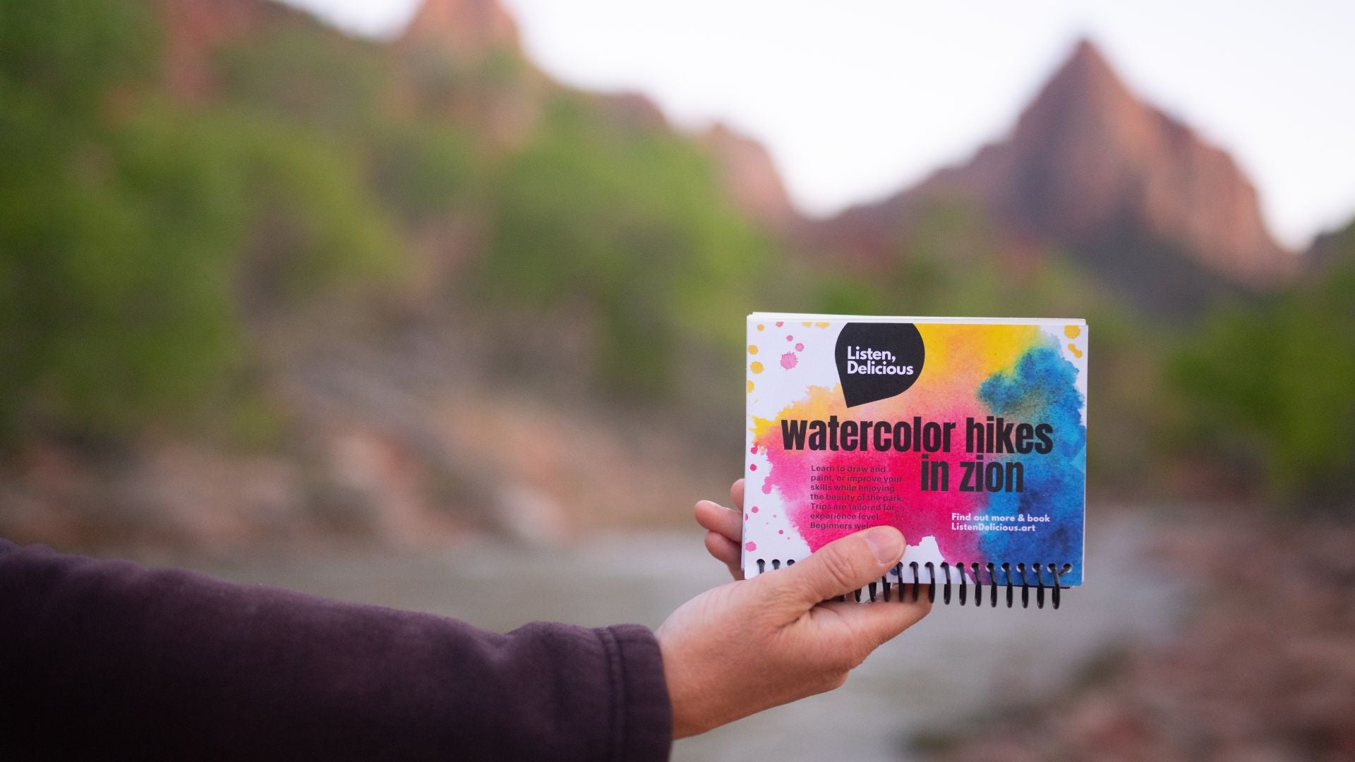 a hand holding a postcard that says watercolor hikes with Zion National Park in the background